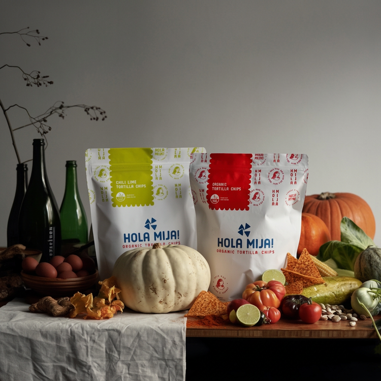 Hola Mija Chili Lime and Organic tortilla chip bags on a harvest table with pumpkins, limes and tomatoes